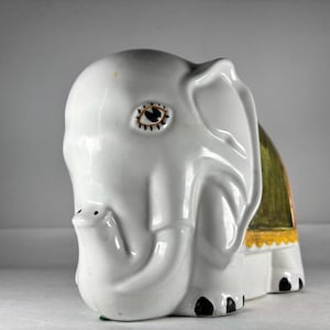 Könnte beinhalten: Weiße Keramik-Elefantenfigur mit einem grün-gelben Sattelmuster. Der Elefant hat ein schwarzes Auge mit einer weißen Pupille und einen kleinen Rüssel.