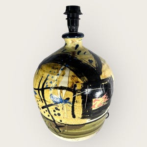 Peut inclure: Une lampe de table en céramique avec un design abstrait en noir, jaune, bleu et blanc. La lampe a une base ronde et un abat-jour noir.