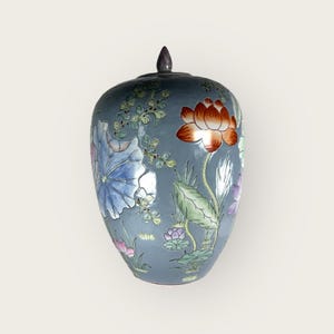 Peut inclure: Un vase en céramique bleu avec un motif floral. Le vase est décoré de fleurs roses, bleues, vertes et orange. Le vase a un couvercle avec un bouton sur le dessus.