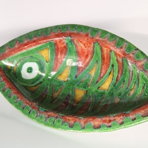 Puede incluir: Un plato de cerámica verde y rojo con forma de pez con un ojo amarillo y blanco. El plato tiene un borde festoneado.
