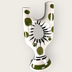 Peut inclure: Un vase en céramique blanc avec un design unique en forme de U. Le vase présente des pois verts et des accents linéaires noirs autour d'une ouverture centrale. La base a des rayures vertes et noires. Un objet décoratif pour la maison.