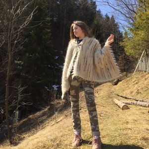 May include: A beige and brown fringed poncho with vertical stripes. The poncho is worn over camouflage pants and pink sneakers. The background features a forest and a sunny day.