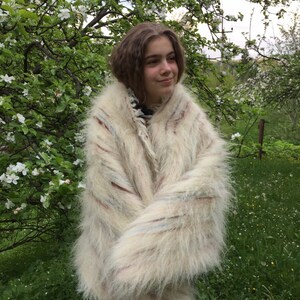 May include: A white, fluffy, striped mohair sweater with a long, loose fit. The sweater is worn by a person standing in a field of green grass and white flowers.