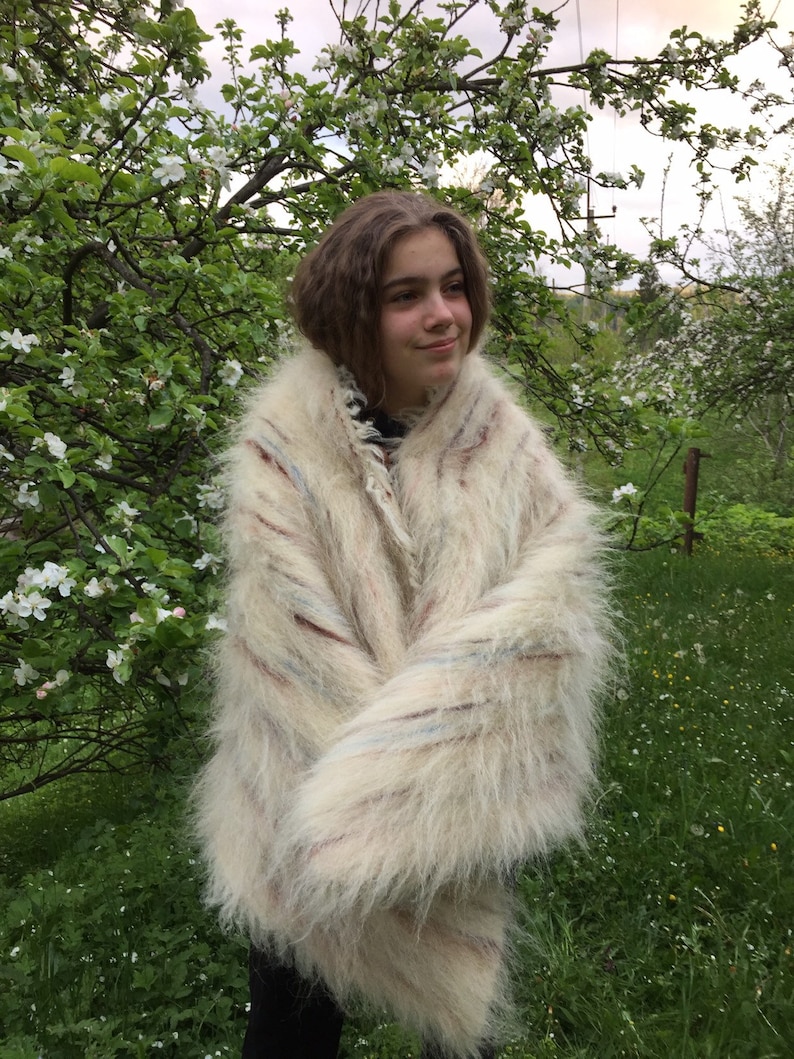 May include: A white, fluffy, striped mohair sweater with a long, loose fit. The sweater is worn by a person standing in a field of green grass and white flowers.