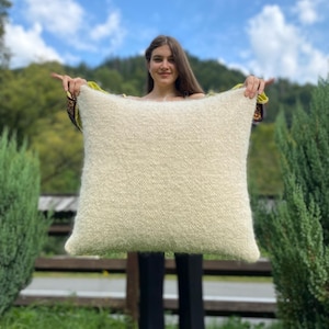 May include: A large, fluffy white pillow held by a person in front of a green background.