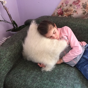 May include: A young person is sleeping on a green couch with a white fluffy pillow. The couch has a fringe trim. The person is wearing a pink long-sleeved shirt and blue jeans.