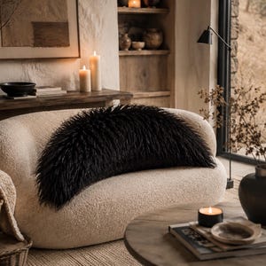 May include: A curved, cream-colored boucl&eacute; sofa featuring a long, rectangular black faux fur throw pillow placed horizontally across the center of the seat. The sofa has a rounded backrest and a low-profile design with a textured fabric finish.