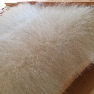 May include: A white fluffy faux fur throw blanket with long, soft fibers. The blanket is draped over a wooden surface.