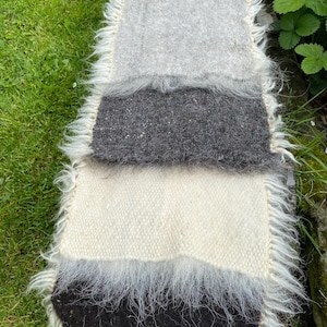 May include: A rectangular rug with a shaggy border, featuring bands of grey, dark grey, white, and black. The rug's texture suggests woven wool or a similar material, giving it a rustic appearance. The fringed edges add to the rug's texture.
