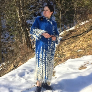 May include: A woman wearing a long, blue and white fuzzy coat stands in a snowy landscape. The coat has a long, flowing design with white accents.