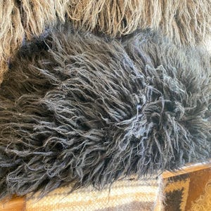 May include: A plush, charcoal black sheepskin pillow featuring long, shaggy, and textured fur fibers. The dense, dark material creates a voluminous appearance with a natural, wild aesthetic. The pillow is positioned in front of a tan fur texture.