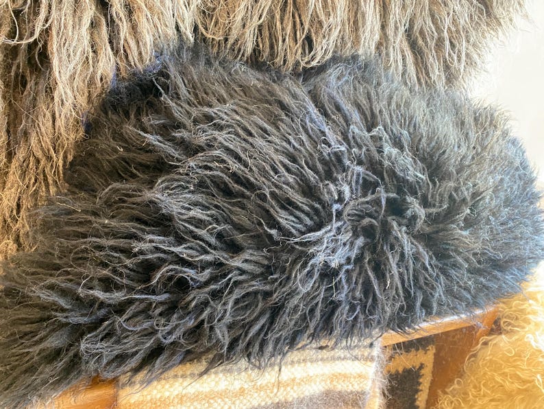 May include: A plush, charcoal black sheepskin pillow featuring long, shaggy, and textured fur fibers. The dense, dark material creates a voluminous appearance with a natural, wild aesthetic. The pillow is positioned in front of a tan fur texture.