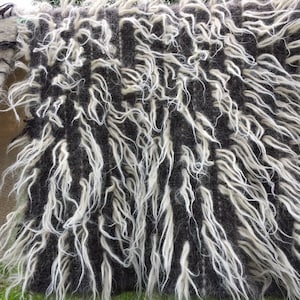 May include: Close-up of a textured textile with a shaggy, layered design. The main section features a dark gray base with long, white, wavy fibers. Additional sections show striped and fringed patterns in shades of brown and cream, suggesting a woven or knitted craft.