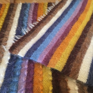 May include: A close-up of two woven rugs with colourful stripes in shades of brown, white, yellow, orange, purple, and blue. The rugs are made of fuzzy yarn and have a textured surface.