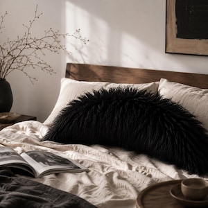 May include: A semi-circular black faux fur pillow with long, shaggy fibers positioned on a bed with light beige linen bedding. The pillow has a distinct curved shape and a dense, textured appearance, contrasting with the smooth fabric of the bed.