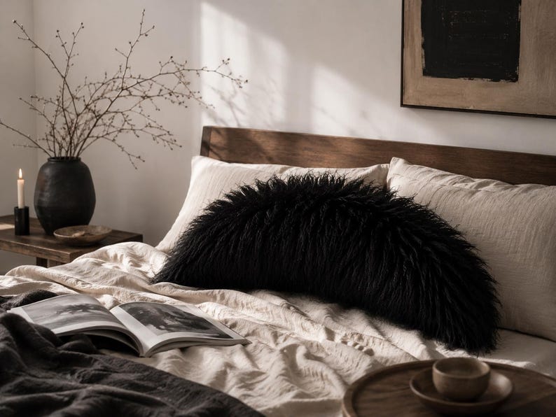May include: A semi-circular black faux fur pillow with long, shaggy fibers positioned on a bed with light beige linen bedding. The pillow has a distinct curved shape and a dense, textured appearance, contrasting with the smooth fabric of the bed.