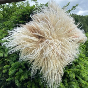 May include: A fluffy white sheepskin rug with a light brown patch in the center. The rug is hanging from a wooden beam and is surrounded by green foliage.