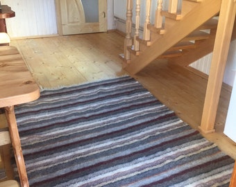 Puede incluir: Una alfombra rayada con rayas marrones, grises y blancas está colocada sobre un suelo de madera. La alfombra está delante de una escalera de madera con una barandilla de madera.