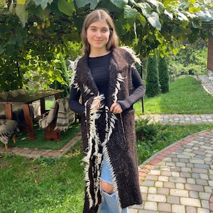 Könnte beinhalten: Eine lange braune Strickweste mit weißen Fransen. Die Weste wird über einem schwarzen Langarmshirt und einer blauen Jeans getragen.