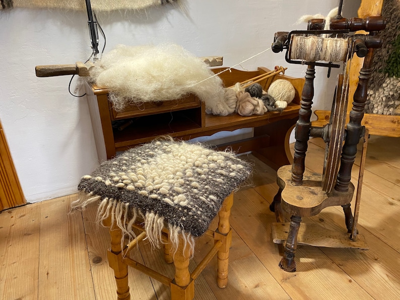 Pode incluir: Uma roda de fiar de madeira com um fio de l&atilde; branco no fuso. A roda est&aacute; sobre uma base de madeira com um pedal. H&aacute; um monte de fibra de l&atilde; branca sobre uma mesa de madeira no fundo.