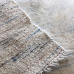 May include: A close-up of a white woven rug with blue and brown stripes. The rug has a fuzzy texture and a frayed edge.