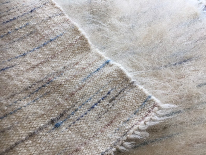 May include: A close-up of a white woven rug with blue and brown stripes. The rug has a fuzzy texture and a frayed edge.