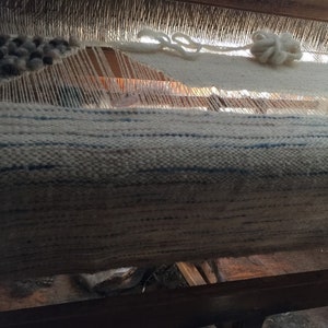 May include: A close-up of a loom with a white and blue woven fabric. The fabric is being woven on a wooden loom with a white yarn hanging from the top.