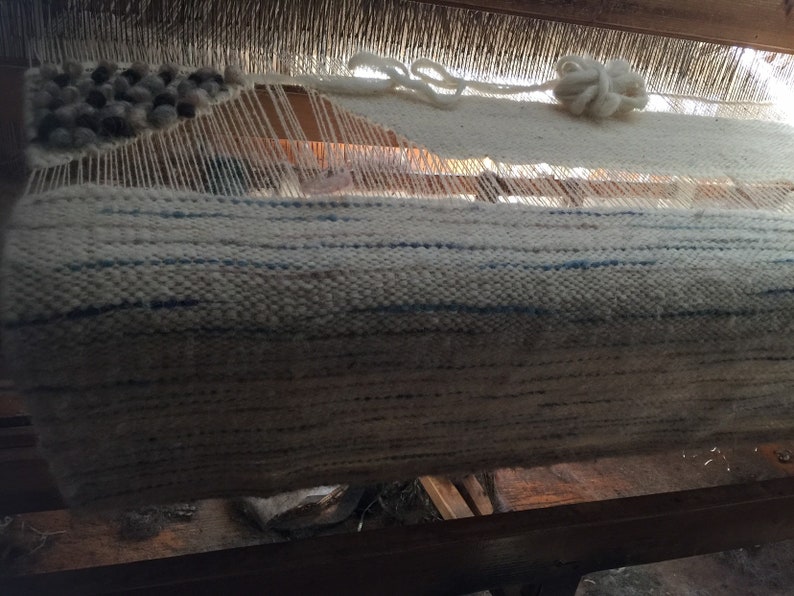 May include: A close-up of a loom with a white and blue woven fabric. The fabric is being woven on a wooden loom with a white yarn hanging from the top.