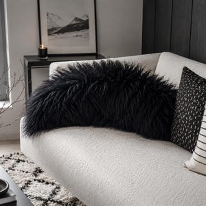 May include: A long, black, shaggy faux fur bolster pillow resting on a white textured sofa. The pillow features a dense, long-pile texture that creates a rounded, voluminous shape across the center of the couch, contrasting with the white fabric.