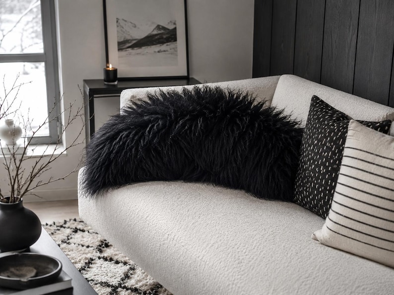 May include: A long, black, shaggy faux fur bolster pillow resting on a white textured sofa. The pillow features a dense, long-pile texture that creates a rounded, voluminous shape across the center of the couch, contrasting with the white fabric.
