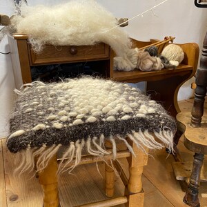 Pode incluir: Um banquinho de madeira com um assento de l&atilde; marrom e branco. O assento tem uma franja ao redor das bordas. O banquinho est&aacute; em uma sala com uma roda de fiar e outros itens de l&atilde;.