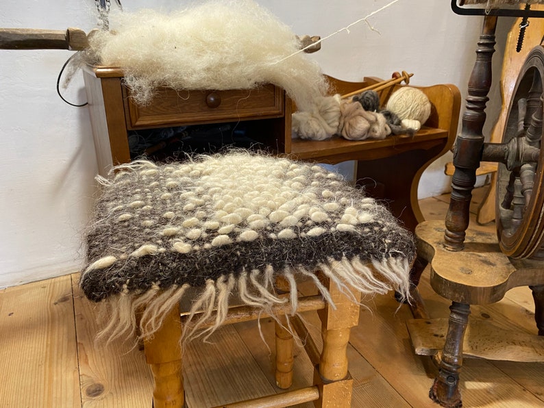 Pode incluir: Um banquinho de madeira com um assento de l&atilde; marrom e branco. O assento tem uma franja ao redor das bordas. O banquinho est&aacute; em uma sala com uma roda de fiar e outros itens de l&atilde;.