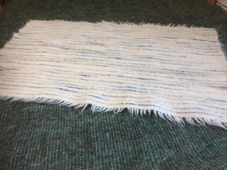 May include: A white woven rug with blue and gray stripes. The rug has a fringed edge and is made of a soft, fluffy material.