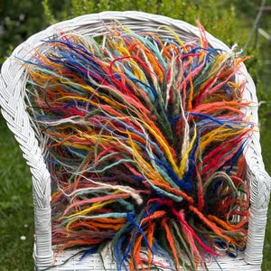 May include: A white wicker chair holds a vibrant, multi-colored fiber art piece. The art features long, textured strands in shades of red, orange, yellow, blue, and green. The chair is set outdoors against a backdrop of green foliage.