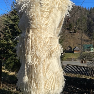 Könnte beinhalten: Cremefarbene, handgefertigte Weste mit zotteligem, strukturiertem Design. Die Weste hat einen gewebten Körper und eine Fransenborte mit einem Kordelverschluss. Das Kleidungsstück wird im Freien auf einer Schaufensterpuppe mit natürlichem Hintergrund präsentiert.