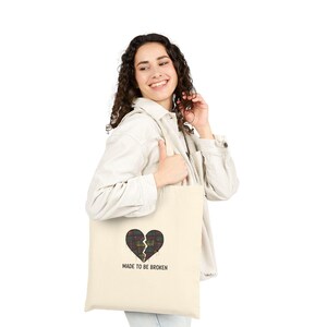 Könnte beinhalten: Cremefarbene Tragetasche mit einem Design eines zerbrochenen Herzens in einem Karomuster. Der Text "MADE TO BE BROKEN" ist unter dem Herzen aufgedruckt. Die Tasche wird von einer Person gehalten.