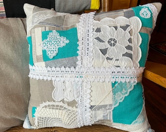 May include: A square pillow with a patchwork design featuring white lace and turquoise fabric. The pillow is made from repurposed materials and has a rustic, vintage aesthetic.