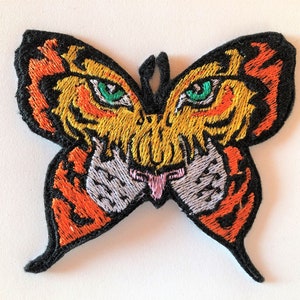 Op de afbeelding: Een geborduurde strijkpatch van een vlinder met een tijgergezicht. De vlinder is zwart met oranje en gele strepen. Het tijgergezicht heeft groene ogen en een roze tong.