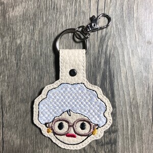 Könnte beinhalten: Ein beigefarbener Leder-Schlüsselanhänger mit einem silbernen Schlüsselring und einem gestickten Bild einer Frau mit Brille in Weiß und Blau.