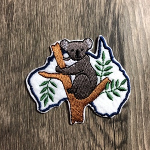 May include: Embroidered patch of a koala bear sitting on a tree branch in the shape of the outline of Australia. The koala is brown and gray, the tree branch is brown, and the leaves are green.
