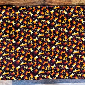 May include: A rectangular, quilted fabric item with a repeating pattern of dachshund dog silhouettes and bone shapes in shades of gold and orange on a dark brown background. The fabric is folded over at the top.
