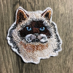 Puede incluir: Parche bordado de un gato blanco y marrón con ojos azules. El gato mira directamente al espectador con una expresión seria.