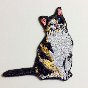 May include: An embroidered iron-on patch of a calico cat sitting with a black, white, and gold pattern. The cat has blue eyes and a pink nose.