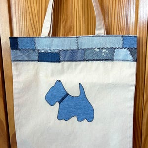 May include: Cream-colored tote bag with a blue denim patchwork top section and a blue dog applique. The bag has cream-colored handles and is made of a sturdy fabric. The dog applique is a stylized Scottish Terrier.