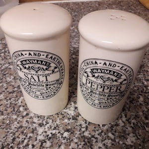 Puede incluir: Dos saleros y pimenteros de cerámica blanca con letras negras. El salero dice "Waymans 60 Redcliffe Street Salt Genuine Finest Quality Made in England". El pimentero dice "Waymans 60 Redcliffe Street Pepper Genuine Finest Quality Made in England".