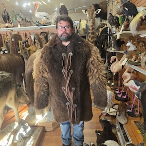 May include: A brown fur coat with leather closures. The coat is knee-length and has a shaggy texture. The person is wearing blue jeans and black shoes. The background features taxidermied animals.