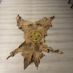 May include: A brown leather hide with painted designs. The center of the hide features a yellow circle with blue and red accents. The hide also features painted images of birds and buffalo.