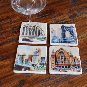 May include: Four square coasters with images of different landmarks in Cleveland, Ohio. The coasters are made of white marble with a smooth, polished surface. The images are printed in vibrant colors and feature the Rock and Roll Hall of Fame, the Terminal Tower, the Hope Memorial Bridge, and the Playhouse Square.