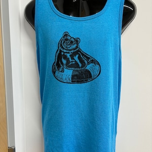 Può includere: Una canotta blu con una grafica nera di un orso che indossa occhiali da sole e galleggia su un galleggiante per piscina.