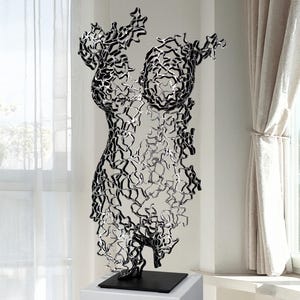 Op de afbeelding: Een zwart-zilveren metalen sculptuur van een vrouwelijk torso. De sculptuur is gemaakt van onderling verbonden, abstracte vormen en staat op een witte sokkel met een zwarte basis. De sculptuur staat bij een raam met transparante gordijnen.